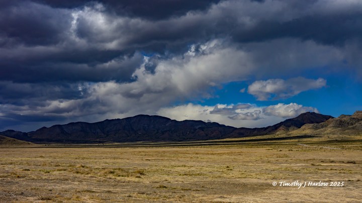 A vast desert landscape under a cloudy sky, featuring distant mountains and dry, sparse vegetation.