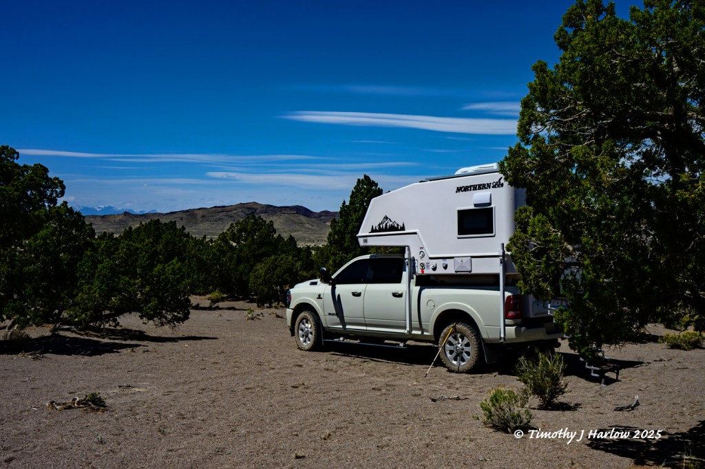 A truck with a camper is parked in a desert landscape, surrounded by sparse vegetation and under a blue sky with clouds.